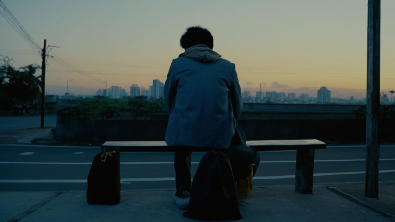 A young person in a blue jacket seen from behind, sitting on a bench at dusk with two bags beside them, a city skyline on the horizon (AI-made image)